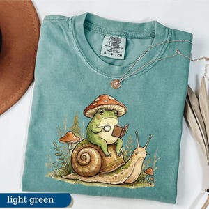 May include: Light green t-shirt with a whimsical illustration of a frog wearing a mushroom hat, reading a book, and holding a cup of coffee while riding a snail. The shirt has a Comfort Colors tag. A necklace is draped on the shirt.