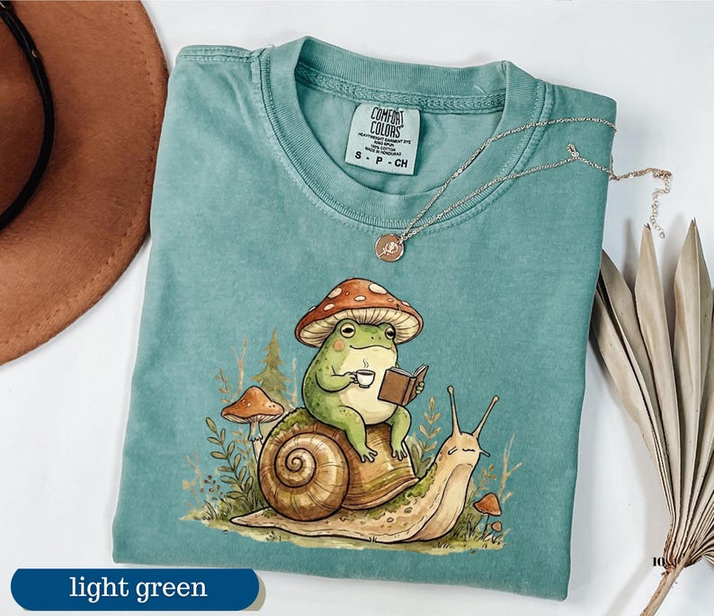 May include: Light green t-shirt with a whimsical illustration of a frog wearing a mushroom hat, reading a book, and holding a cup of coffee while riding a snail. The shirt has a Comfort Colors tag. A necklace is draped on the shirt.