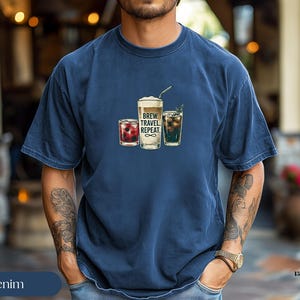 Puede incluir: Camiseta azul marino con un gr&aacute;fico de tres bebidas: una bebida de bayas, una bebida de caf&eacute; con pajita y una bebida verde. El texto "BREW. TRAVEL. REPEAT." est&aacute; impreso.
