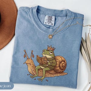 May include: A blue jean colored t-shirt with a whimsical illustration of a frog wearing a crown and sweater, reading a book titled "Tales of Cottage". A snail is also featured. A gold necklace is draped across the shirt.