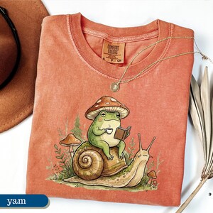 May include: A coral-colored Comfort Colors t-shirt featuring a whimsical illustration of a frog wearing a mushroom hat, reading a book, and drinking coffee while riding a snail. A gold necklace and a brown hat are also visible.