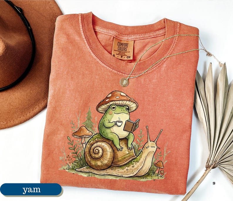 May include: A coral-colored Comfort Colors t-shirt featuring a whimsical illustration of a frog wearing a mushroom hat, reading a book, and drinking coffee while riding a snail. A gold necklace and a brown hat are also visible.