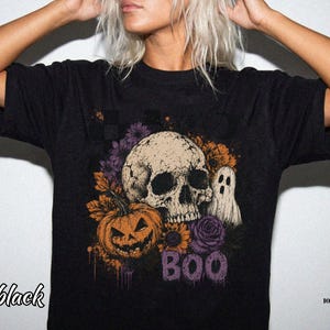 May include: A black t-shirt featuring a graphic print of a beige human skull, a carved orange jack-o'-lantern, a white ghost, and purple and orange flowers. The design includes a checkered pattern and the word BOO in purple letters.