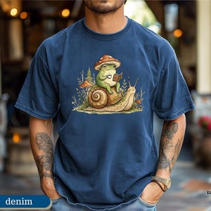 May include: Denim blue t-shirt featuring a whimsical illustration. A green frog wearing a mushroom hat is reading a book while riding on a snail. The design includes mushrooms and foliage, creating a nature-inspired theme.