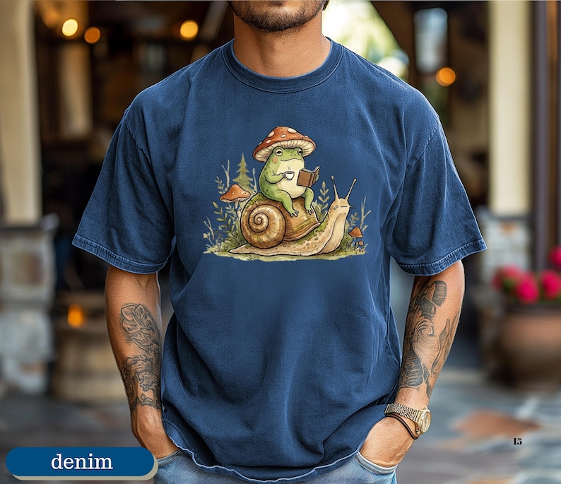 May include: Denim blue t-shirt featuring a whimsical illustration. A green frog wearing a mushroom hat is reading a book while riding on a snail. The design includes mushrooms and foliage, creating a nature-inspired theme.