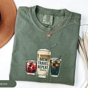 Puede incluir: Camiseta verde musgo Comfort Colors con un gr&aacute;fico de tres vasos de bebidas. El vaso central tiene el texto "BREW. TRAVEL. REPEAT." La camiseta tiene cuello redondo y mangas cortas. Una cadena de oro est&aacute; en la camiseta.