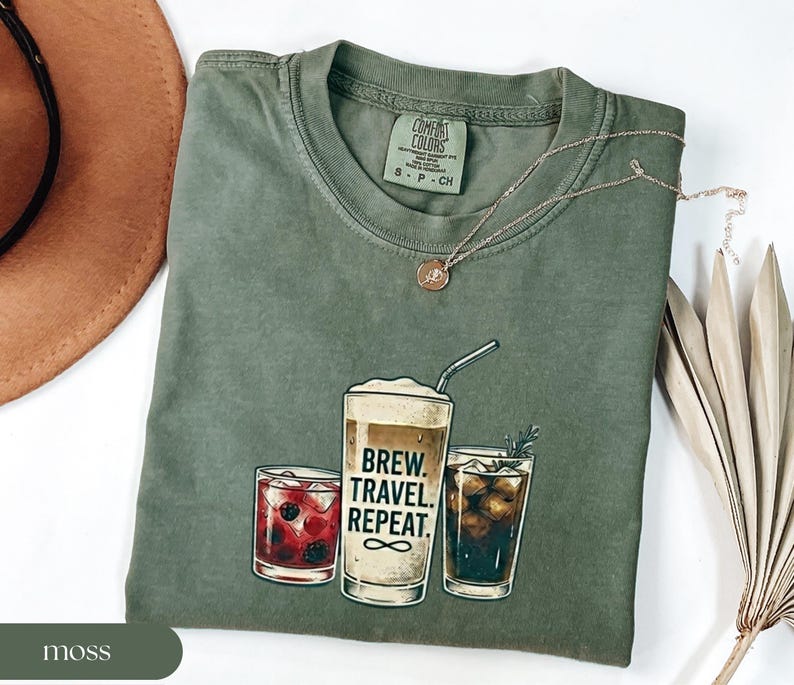 Puede incluir: Camiseta verde musgo Comfort Colors con un gr&aacute;fico de tres vasos de bebidas. El vaso central tiene el texto "BREW. TRAVEL. REPEAT." La camiseta tiene cuello redondo y mangas cortas. Una cadena de oro est&aacute; en la camiseta.