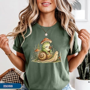 May include: Moss green t-shirt featuring a whimsical illustration of a frog wearing a mushroom hat, reading a book, and riding a snail. The frog is holding a cup. The design includes other mushrooms and plants.