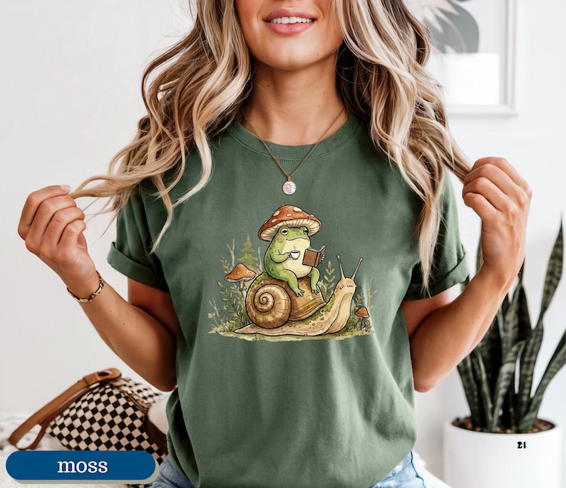 May include: Moss green t-shirt featuring a whimsical illustration of a frog wearing a mushroom hat, reading a book, and riding a snail. The frog is holding a cup. The design includes other mushrooms and plants.