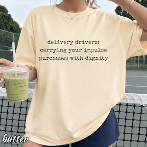 May include: A butter-colored short-sleeve t-shirt featuring black typewriter-style text that reads: delivery drivers: carrying your impulse purchases with dignity. The shirt has a crew neckline and a loose, relaxed fit, made of cotton fabric.