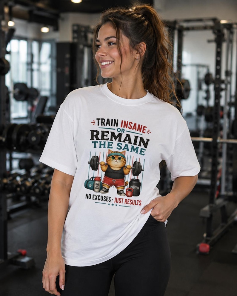 May include: A white short-sleeve t-shirt featuring a graphic of a cat lifting a barbell with the text "TRAIN INSANE OR REMAIN THE SAME" and "NO EXCUSES - JUST RESULTS". The cat wears a headband, tank top, and shorts, with a gym bag and protein tub.