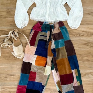 May include: An overhead shot of a fashion ensemble featuring a white, long-sleeved blouse with floral embroidery, patchwork corduroy pants in various colors, and beige espadrille sandals with tie-up straps. The outfit is laid out on a wooden surface.