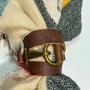 May include: A brown leather scarf ring with a brass buckle secures a colorful scarf. The scarf features a plaid pattern in cream, teal, yellow, gray, and black. The leather ring is a stylish accessory.