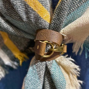 May include: A brown leather scarf slide featuring a brass-toned metal buckle closure. The leather band is wrapped around a woven scarf that displays a pattern of yellow, teal, and cream-colored stripes with frayed edges on the fabric ends.