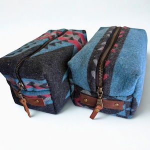 May include: Two rectangular zippered pouches featuring a woven geometric pattern in shades of blue, charcoal gray, and red. Each bag has a dark metal zipper closure with a brown leather pull tab attached by two brass rivets at the front end.