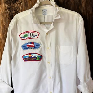 May include: A white long-sleeve button-down shirt with a ruffled collar and three embroidered patches on the right chest. The patches feature text including Three Rivers Council, Indianhead Council, and Denver Area Council Colorado.