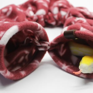 May include: A burgundy and white patterned fleece tube with a brown leather accent. A yellow and white toy snake with an orange tongue is inside the tube. The tube is designed for small pets.