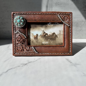 May include: Brown leather-look picture frame with a turquoise stone accent and decorative stitching. The frame holds a 6"x4" image of horses and cowboys. The frame has a western theme.