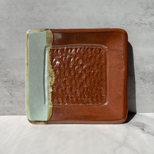May include: A square ceramic dish featuring a glossy, deep reddish-brown glaze on the main surface with a textured, repeating pattern. One side of the dish is finished with a smooth, contrasting pale mint green glaze and a tan border.