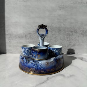 May include: A circular ceramic condiment server featuring a central handle and four small cups. The base and cups are decorated with a deep blue and white floral pattern, accented with gold gilding along the edges and textured surfaces.