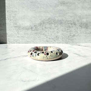 Puede incluir: Un anillo de cerámica moteada con una serie de agujeros espaciados uniformemente. El anillo presenta una base de color crema con acentos de morado, azul y verde. El objeto es circular y se encuentra sobre una superficie blanca.