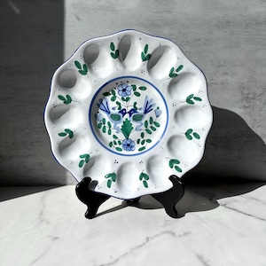 May include: White ceramic egg plate with a scalloped edge and a blue rim. The plate features a central floral design in blue and green, surrounded by individual egg compartments. Green leaf accents are scattered around the edge. The plate is displayed on a black stand.