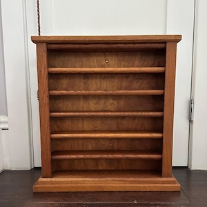 May include: A rectangular wooden wall-mounted display shelf featuring a medium brown oak finish with a visible wood grain. The unit consists of five horizontal shelves, each fronted by a rounded wooden rail, and a solid back panel with two small holes.