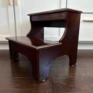 May include: A dark brown wooden two-step stool with a smooth, polished finish. The stool features a rectangular top step and a lower step, supported by solid side panels with arched cutouts at the base, providing a stable structure.