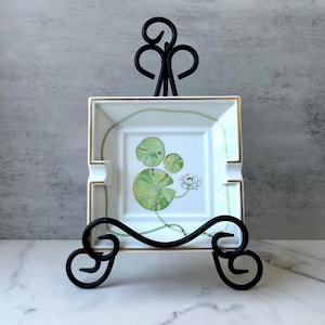 May include: Square white ashtray with a hand-painted water lily design, featuring green lily pads and a white flower. The ashtray has gold trim and is displayed on a black metal stand with decorative swirls.