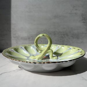 May include: A round, pale green ceramic serving dish with a central loop handle. The dish features twelve individual indentations for eggs, decorated with delicate floral patterns and gold-painted scalloped edges along the rim and handle.