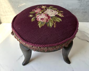May include: A small, round footstool with a dark burgundy fabric top featuring a floral needlepoint design in pink, white, and green. The edge is trimmed with a twisted rope and decorative trim. The stool has dark wooden legs.