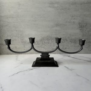 May include: A dark grey metal candelabra with four candle holders. The candelabra has a square base and curved arms. The candle holders are small and cylindrical. The background is a light grey wall and a white marble surface.