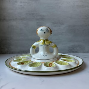 May include: A vintage ceramic deviled egg serving dish. The white dish has a circular design with a central figurine and egg-shaped indentations. The figurine has a yellow collar and brown hair. The dish has green and orange accents.