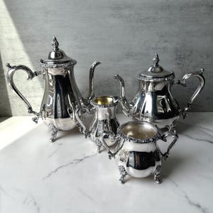 May include: A silver tea set comprising a teapot, a creamer, a sugar bowl, and a coffee pot. The set features ornate handles and decorative trim. The items are arranged on a marble surface.