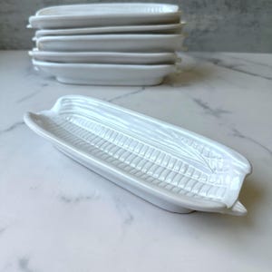 May include: White ceramic corn-shaped serving dishes. The dishes have a detailed design mimicking corn on the cob. A stack of similar dishes is visible in the background. The dishes are on a marble surface.