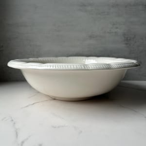 May include: A white ceramic bowl with a scalloped rim and a textured, beaded edge. The bowl is set against a neutral background, highlighting its simple elegance. This bowl is suitable for serving or display.