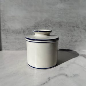 May include: A cylindrical ceramic butter crock with a smooth off-white finish and a matching lid. The piece features thin, dark blue horizontal bands encircling the top edge of the lid, the base of the lid, and the bottom of the main container.