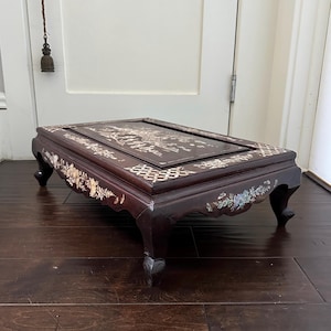 May include: A low rectangular dark wood table featuring intricate mother-of-pearl inlay designs. The surface displays a detailed landscape scene, while the edges and four curved legs are decorated with floral patterns and geometric lattice motifs.