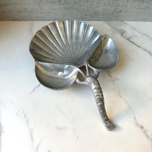 Könnte beinhalten: Eine silberfarbene Metallschale in Form einer großen Jakobsmuschel mit zwei kleineren blattförmigen Seitenfächern und einem langen, strukturierten Griff an der Basis, mit metallischem Finish und einer glatten, reflektierenden Oberfläche.