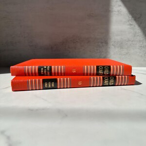 May include: Two bright orange hardcover books stacked horizontally. The spines feature black rectangular labels with gold text reading Childcraft, Your Young Child, and Your Child in Today's World, alongside the numbers 13 and 15 and vertical grey stripes.