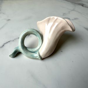 Puede incluir: Un jarrón de cerámica con forma de lirio de agua. El jarrón tiene una flor rosa claro y un tallo rizado azul claro. El jarrón está sobre una superficie blanca.