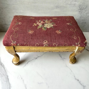 Puede incluir: Un taburete antiguo con una parte superior de tapiz burdeos con un diseño floral en rosa, verde y crema. El marco de madera tiene un acabado dorado envejecido, y las patas curvas añaden un toque de elegancia. Un artículo de decoración vintage.