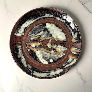以下が含まれることがあります： ユニークな抽象デザインの手作りセラミックプレート。プレートは、濃い茶色の縁と、黒、白、金色のアクセントが施された中央のデザインが特徴です。プレートの表面は質感があり、その職人的な品質を高めています。