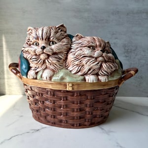 Könnte beinhalten: Ein keramikförmiger Behälter in Form eines Korbes mit zwei flauschigen Katzenfiguren. Die Katzen sind hellbraun mit dunkleren Streifen und sitzen auf einem grünen und blauen Stoff. Der Korb ist braun mit einer gewebten Textur und hat zwei Griffe.