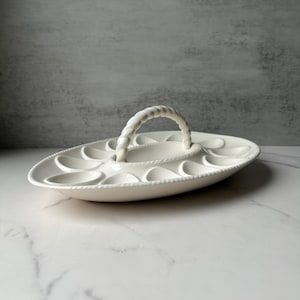 May include: A white ceramic egg serving platter with a woven handle. The oval-shaped dish has multiple egg-shaped indentations around the edge and a central section. The handle is a thick, braided design.