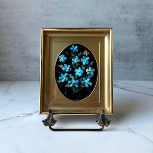 May include: A rectangular gold-toned frame featuring an oval center inlay. Inside the oval, a dark background displays a cluster of small, bright turquoise blue flowers with green stems and leaves, accented by a single small gold-colored butterfly.