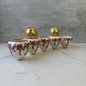 May include: Five white ceramic vases with a heart-like shape, adorned with pink floral patterns and a gold-colored top. The vases are arranged in a row, showcasing a delicate and artistic design.