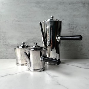 May include: A silver-colored coffee set with a tall coffee pot, a creamer, and a sugar bowl. The coffee pot and creamer have black handles. The set is displayed on a white marble surface.