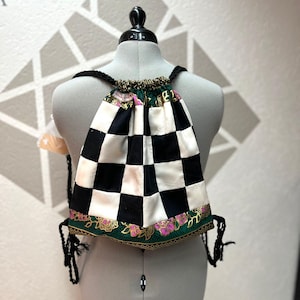 Könnte beinhalten: Ein Rucksack mit Kordelzug und einem schwarz-weißen Schachbrettmuster. Die Tasche hat schwarze geflochtene Träger und eine grüne und rosa geblümte Bordüre. Die Tasche wird auf einer Schaufensterpuppe präsentiert.