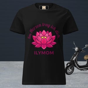Könnte beinhalten: Schwarzes T-Shirt mit einem rosa Lotusblüten-Design und dem Text "Im Herzen trag ich dich. ILYMOM". Das T-Shirt hat einen Rundhalsausschnitt und kurze Ärmel. Ein schwarzer Roller ist im Hintergrund.
